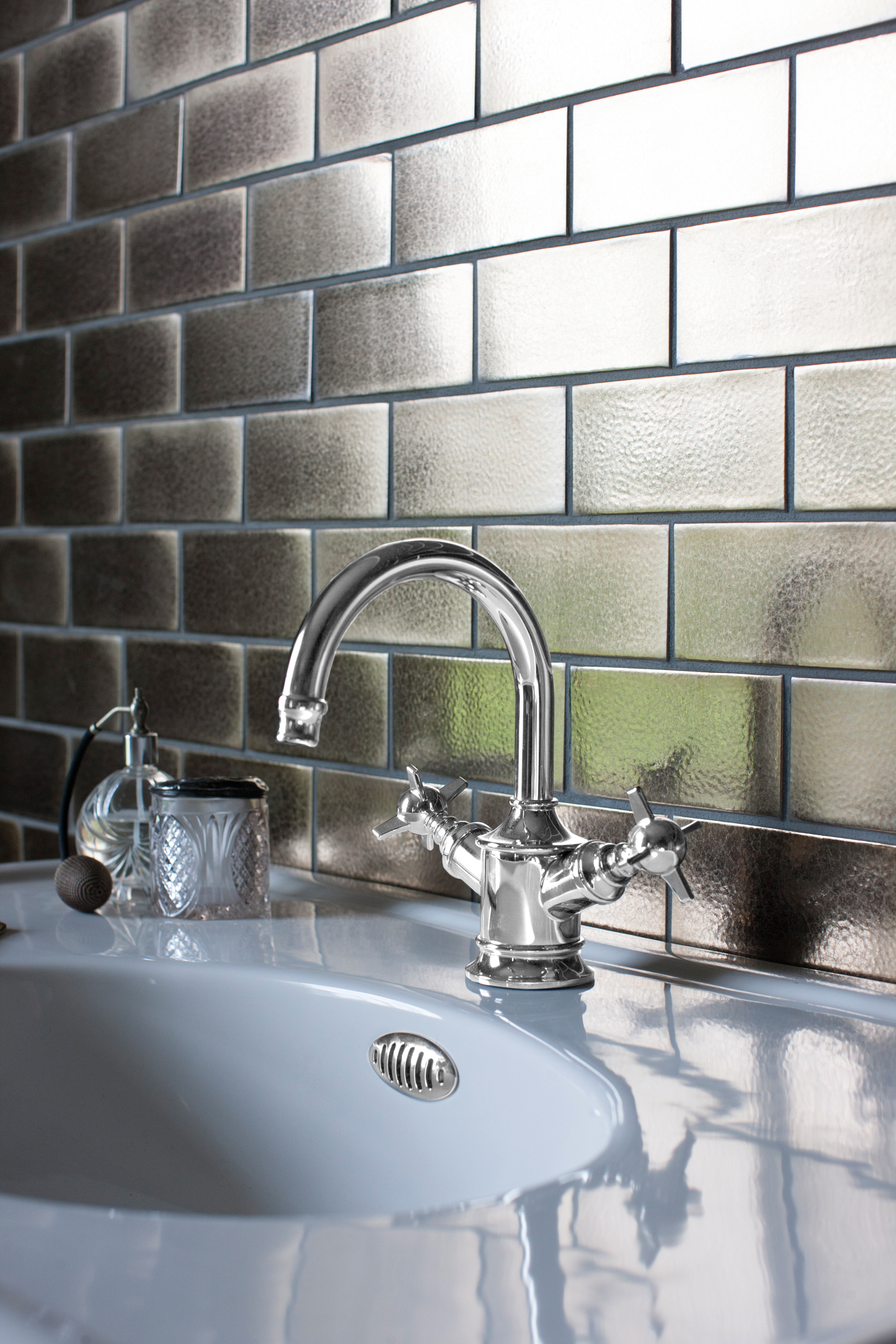 Chrome curved faucet over a white sink with two handles; frosted glass brick wall in the background and small glass jar on the counter.