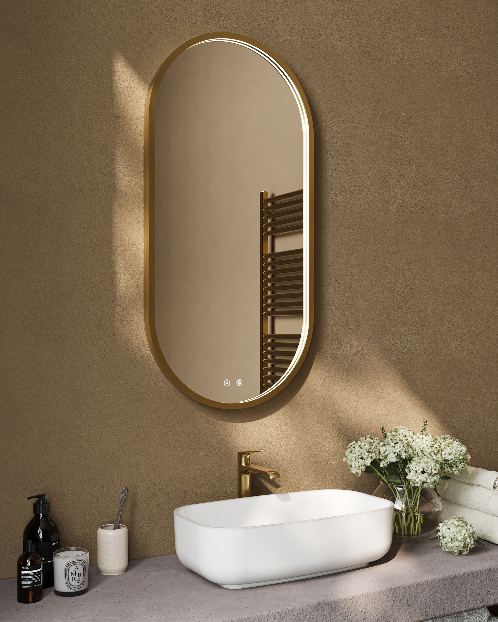 Warm-toned bathroom with a tall oval gold-framed mirror above a white oval sink and gold faucet; a vase of white flowers and toiletries on the counter.