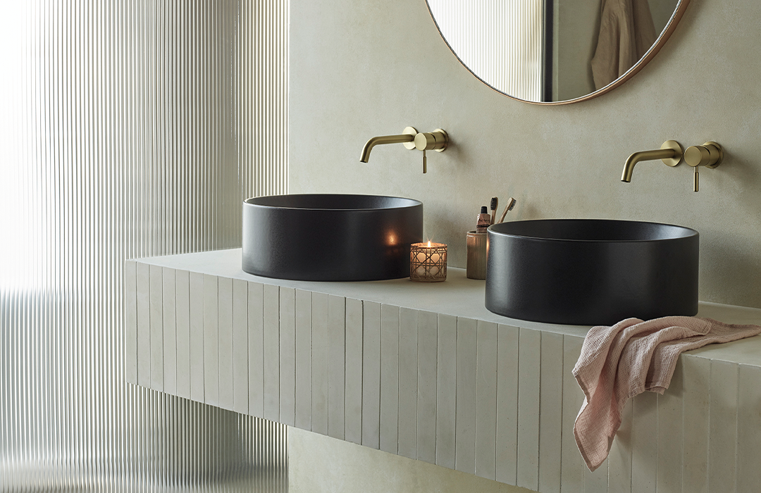 Minimalist bathroom with two matte black round sinks on a light beige countertop, gold wall taps, a round mirror, a lit candle, and a pink towel.