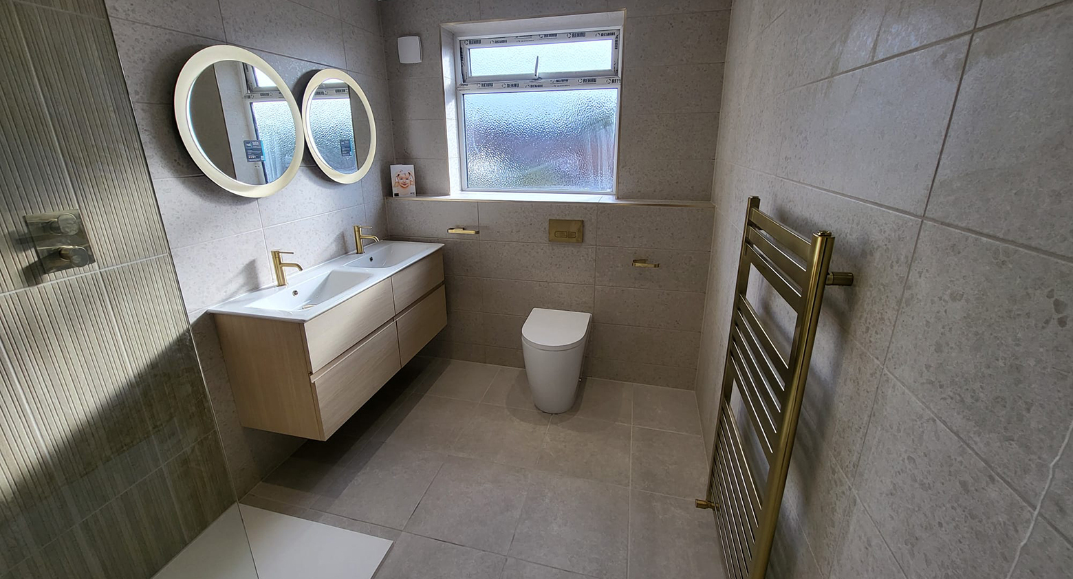 Modern bathroom with a wall-hung wood vanity and double sinks, two round backlit mirrors, frosted window, toilet, and brass heated towel rail.