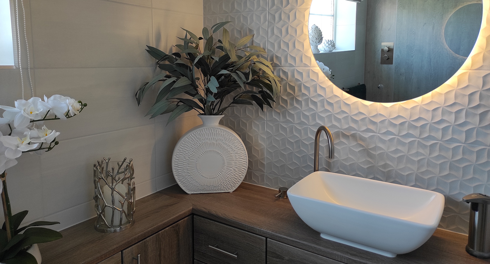 Modern bathroom vanity with a white vessel sink, curved faucet, round backlit mirror, 3D geometric tile wall, and potted plants on a wooden countertop.