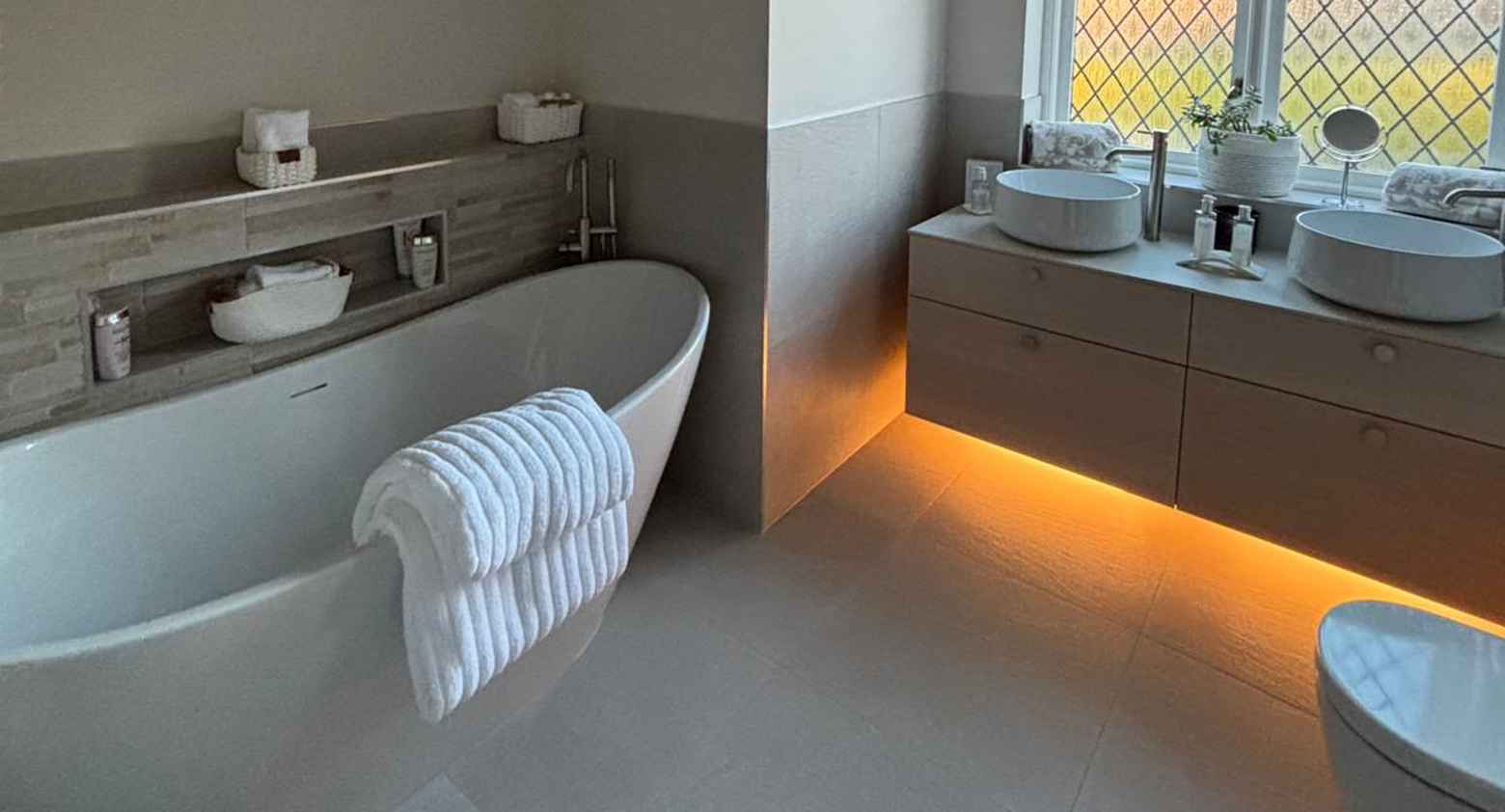 Modern bathroom featuring a freestanding white tub with a fluffy towel over the rim, and a double vanity with vessel sinks beneath warm lighting; diamond-pattern window.
