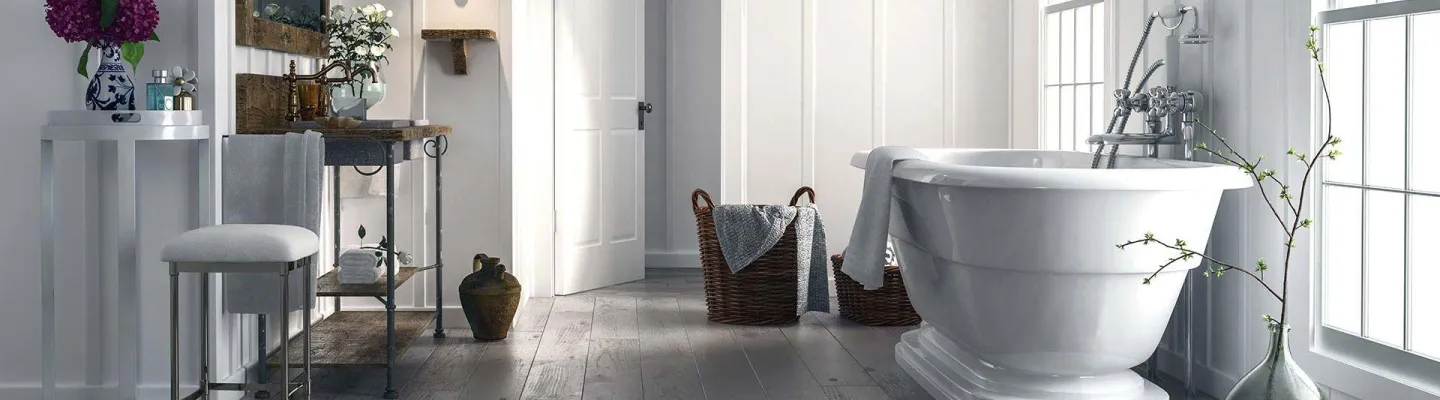 Bright, airy bathroom with a freestanding white tub by a large window, towels on baskets, a wooden vanity, and decorative vases with flowers.