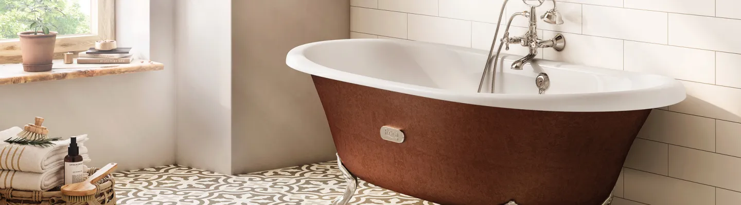 Sunlit bathroom with a vintage brown exterior, white interior clawfoot tub, chrome fittings, white subway tiles, a patterned floor, and a wicker basket of towels.