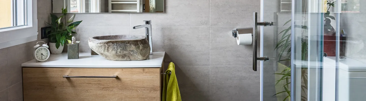 Modern bathroom with grey tiles, a stone vessel sink on a white countertop, wooden vanity, plant, clock, a green towel, and a glass shower enclosure.