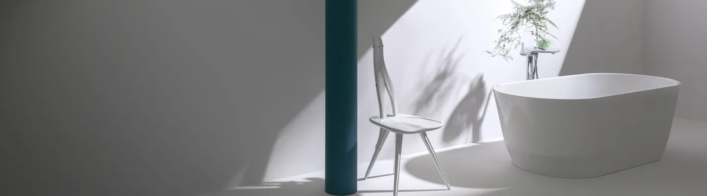 Minimalist bathroom with a white freestanding tub, a white chair beside a teal column, and a potted plant casting shadows on pale walls.