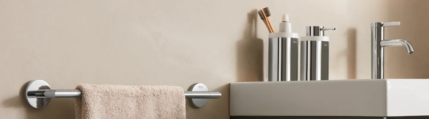 Modern bathroom sink area with beige wall, chrome tap, chrome dispensers, toothbrush holder with brushes, and a beige towel on a chrome rail.