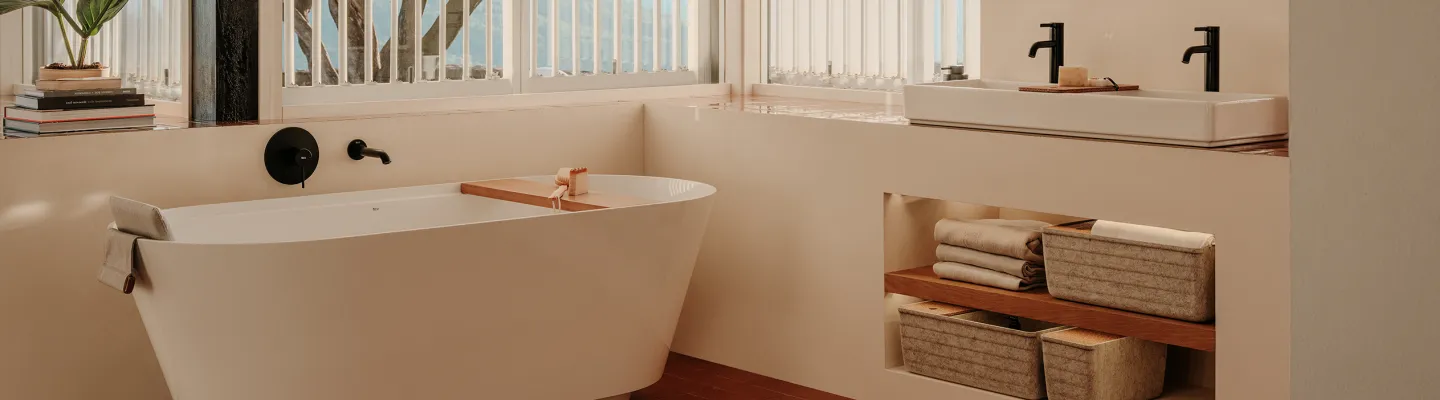 Minimalist bathroom with a white freestanding tub, black fixtures, wooden shelves, folded towels and woven baskets; sunlight through white blinds.