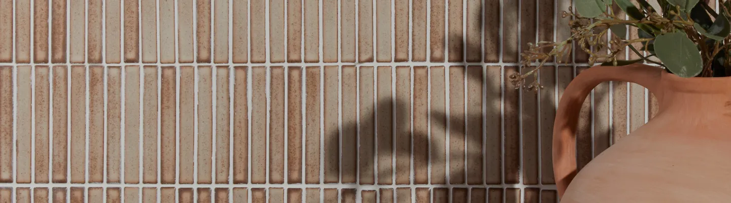 Vertical beige-brown tiled wall with white grout; a terracotta vase with a curved handle sits on the right, holding a green plant.