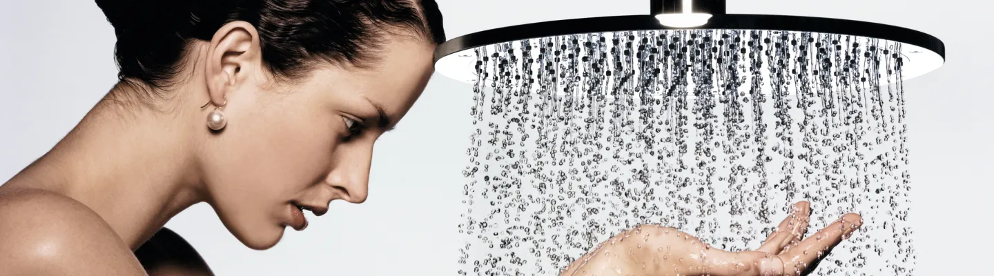 Side profile of a person with a pearl earring, standing under a large rainfall showerhead with water cascading over their hand.