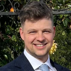Smiling person in a dark suit, white shirt and light blue tie, outdoors with greenery and a wrought-iron fence behind. profile photo