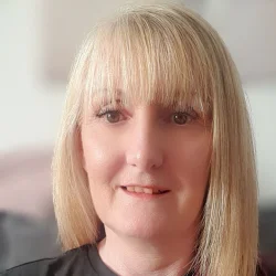 Close-up portrait of a person with straight, shoulder-length blonde hair and bangs, wearing a dark top, neutral expression, light blurred background. profile photo