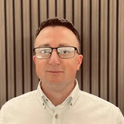 A person with short dark hair and glasses, wearing a light button-up shirt, standing in front of vertical wooden slats with a neutral expression. profile photo