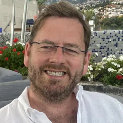 Smiling person with short light brown hair, stubble and glasses, in a white shirt, outdoors by flowers, with a hillside town in the background. profile photo