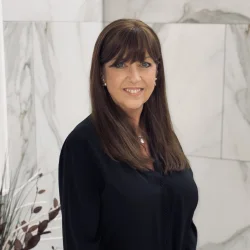 Smiling person with long brown hair and bangs, wearing a black blouse, standing against a white marble wall. profile photo