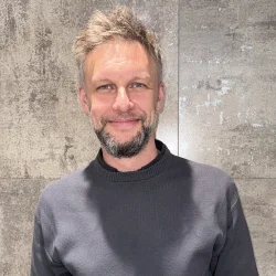 Person with blond hair and a short beard, in a dark grey crew-neck sweater, smiling against a textured grey wall. profile photo