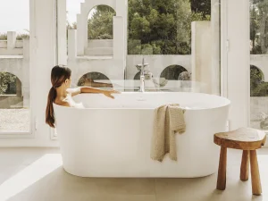 Person sits in a white freestanding bathtub in a bright room with large windows; a beige knitted towel drapes the tub edge, wooden stool nearby, arches outside.