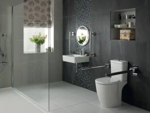 Modern bathroom with glass shower, grey tiles, wall-mounted sink and circular mirror, toilet with grab bars, open shelves, vase of flowers by window.