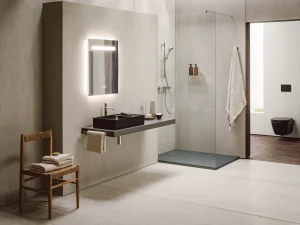 Minimalist bathroom with concrete walls, a backlit mirror, black sink on a floating counter, glass shower and a wooden chair with folded towels.