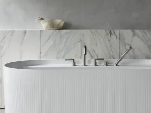 White ribbed freestanding bathtub with chrome fixtures in a marble-tiled bathroom; grey wall above and a woven basket on a ledge.