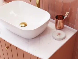 White vessel sink on a pale marble countertop with pink-tiled walls; gold faucet and drain, copper toothbrush holder, and a round marble soap dish.