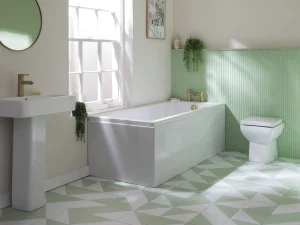 Bright bathroom with a white pedestal sink, freestanding bathtub and toilet, pale green ribbed wall panels, mint and white geometric tiled floor, round mirror, window and plants.