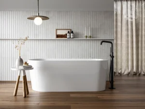 Minimalist bathroom with a white freestanding tub, black floor tap, wood floors, textured white wall, table with dried stems, and beige curtains.