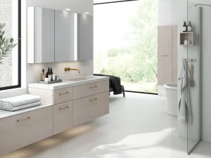 Bright modern bathroom with light-wood vanity, brass tap, large window view of greenery, wall cabinets, glass shower and towel rack.
