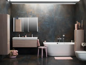 Modern bathroom with dark, metallic walls and a wall-mounted wooden double vanity with two sinks and a backlit mirrored cabinet; a white freestanding tub, pink towels and stool.