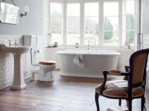 Bright vintage bathroom with a freestanding white bath by a bay window, pedestal sink, toilet with a wooden seat, and ornate dark wood armchair.