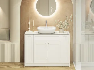 White bathroom vanity with round backlit mirror, vessel sink, and a vase of white blossoms on a beige wall; candles left, glass shower right.