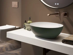 Modern bathroom with a round backlit mirror, white floating counter, green bowl sink, wall-mounted tap, soap bottles, towel, and herringbone floor.