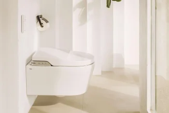 Minimal, bright white bathroom with a wall-mounted toilet, flush plate, and paper holder; pale floor and a hanging green plant by the doorway.