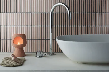 Modern bathroom with oval white basin and chrome curved faucet, terracotta candle holder with a lit candle, woven mat and seashell on countertop.