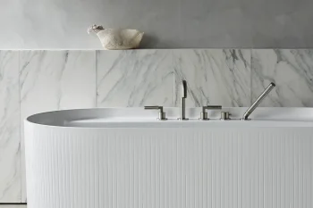 White ribbed freestanding bathtub with chrome fixtures in a marble-tiled bathroom; grey wall above and a woven basket on a ledge.