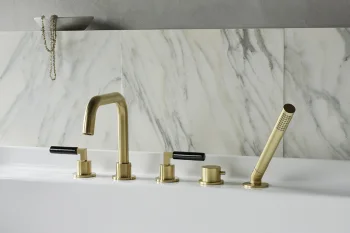 Brass bathroom faucet with black handles and a matching handheld shower, mounted on a white tub with marble backsplash and grey wall.