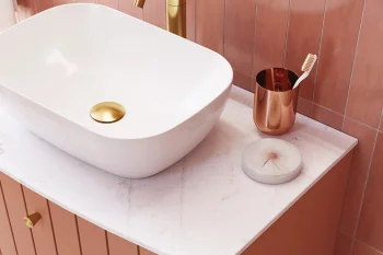 White vessel sink on a pale marble countertop with pink-tiled walls; gold faucet and drain, copper toothbrush holder, and a round marble soap dish.