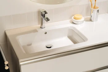 White bathroom sink with chrome faucet, soap on a dish, and a toothbrush holder with two wooden toothbrushes on a glossy white counter.