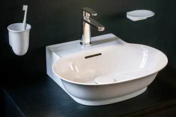 White ceramic sink with chrome tap on a dark wooden vanity, circular backlit mirror above; toothbrush cup left, soap dish right, door reflected.