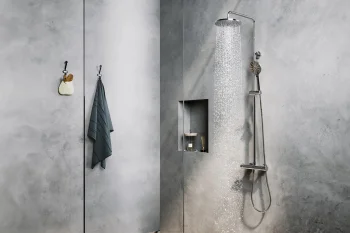 Concrete grey bathroom with a tall rain shower on the right, a handheld unit and hose; a recessed niche holds a candle and diffuser; a towel and small bag hang on the left.
