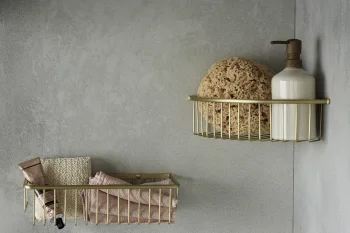 Two brass wire shelves mounted on a grey wall; right shelf holds a pump bottle and a loofah sponge, left shelf contains folded towels and a tube.