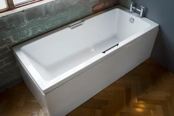 White rectangular bathtub with chrome taps sits against a grey wall next to an exposed brick wall and a window, on a wooden parquet floor.