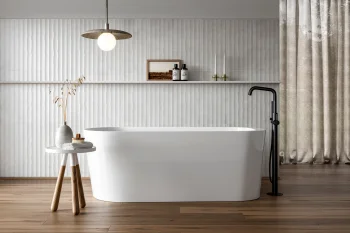 Minimalist bathroom with a white freestanding tub, black floor tap, wood floors, textured white wall, table with dried stems, and beige curtains.