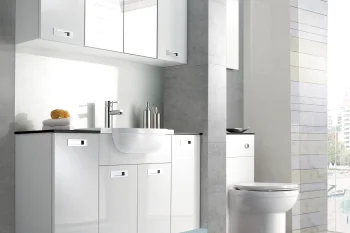 Modern bathroom with white glossy vanity and wall cabinets, chrome faucet, soap dispensers, towel and sponge, toilet beside a window overlooking city.