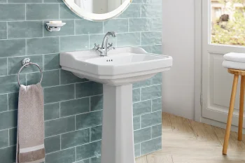 White pedestal sink with chrome taps against teal subway tiles, towel ring with beige towel, wood floor, stool with folded towels by a white door.