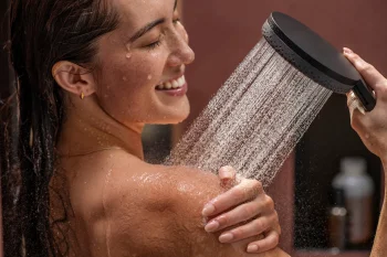 Close-up of a person with wet hair smiling under a handheld shower, water cascading over their shoulder as they wash.