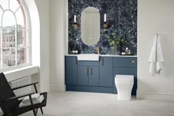 Modern bathroom with dark blue vanity, white inset sink, gold faucet, arched mirror, dark green marble backsplash, two wall sconces, plants, and a white toilet.