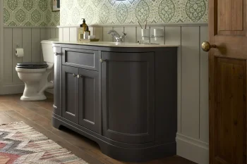 Dark grey curved bathroom vanity with white top and sink, chrome faucet; toiletries, toilet, wood floor, white wainscoting, green damask wallpaper, door.
