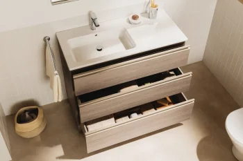 Modern bathroom with a white sink in a light-wood vanity and three open drawers; mirror reflects a toilet and bidet, basket with towels on the floor.
