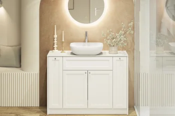 White bathroom vanity with round backlit mirror, vessel sink, and a vase of white blossoms on a beige wall; candles left, glass shower right.